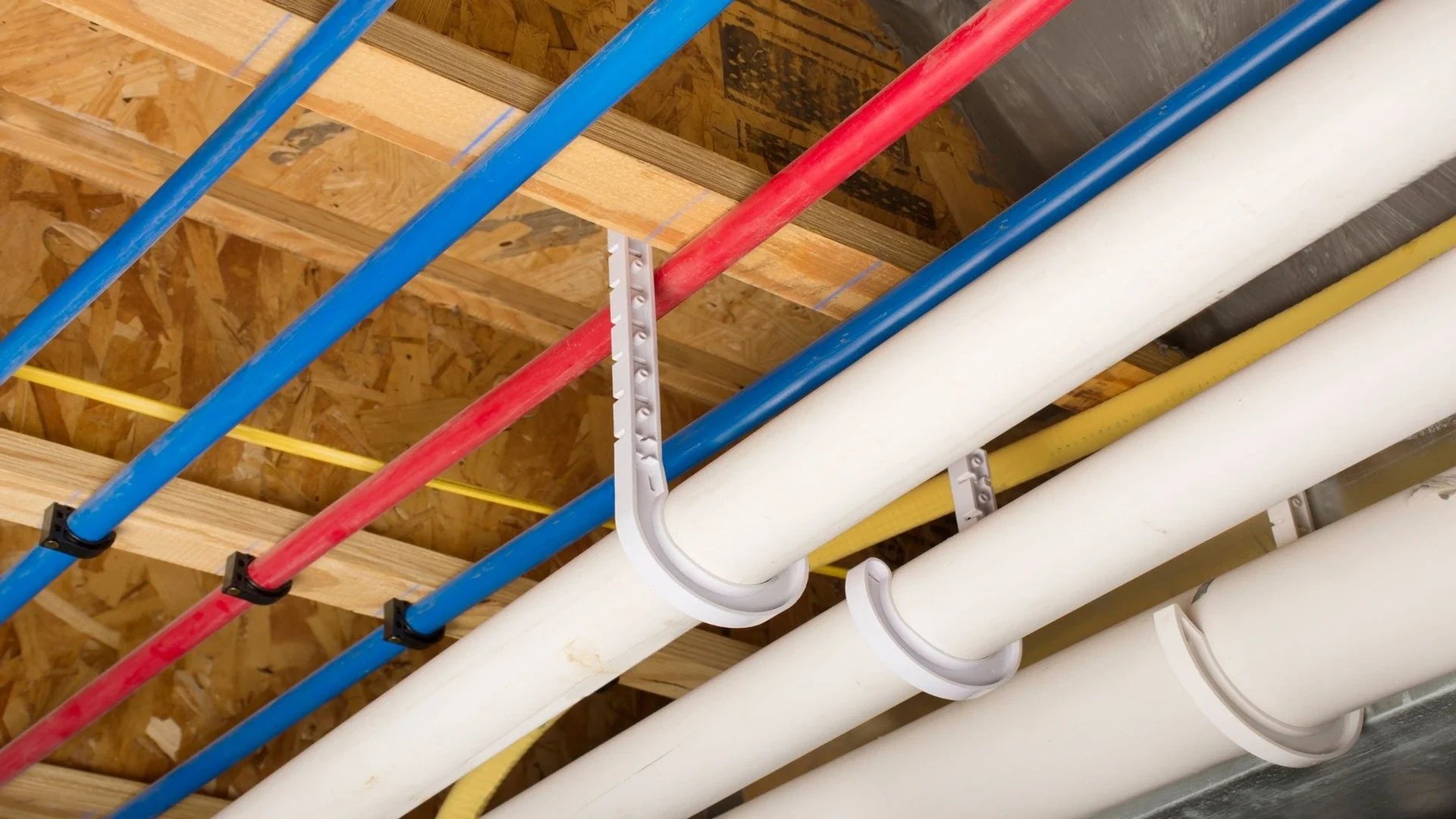 Red, blue, yellow, and white pipes secured with plastic plumbing brackets under wooden ceiling beams.
