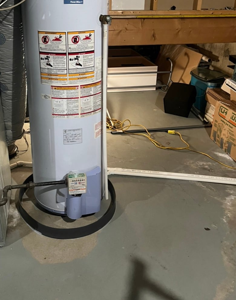 White water heater tank with warning labels installed in basement with concrete floor and stored items.
