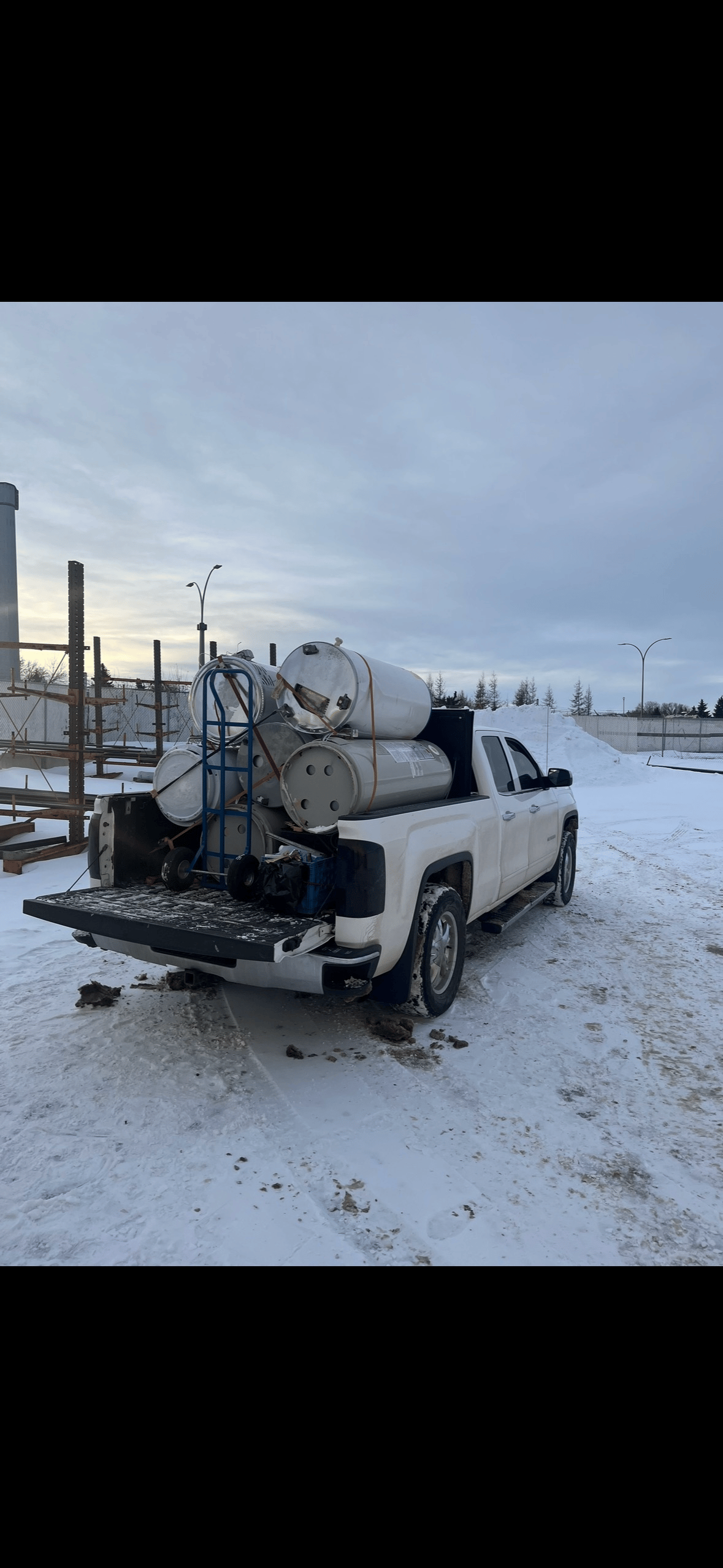 White pickup truck parked in snow with large metal barrels and a hand truck in the bed.