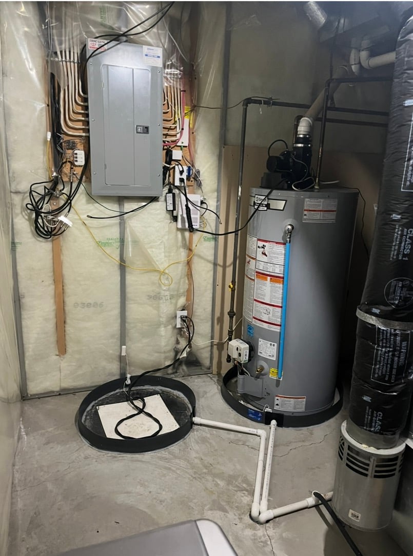 Utility room with electric panel, water heater, insulated walls, and connected pipes on concrete floor.