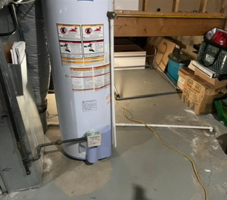 White water heater with pipes in a basement near boxes and storage bins on concrete floor.